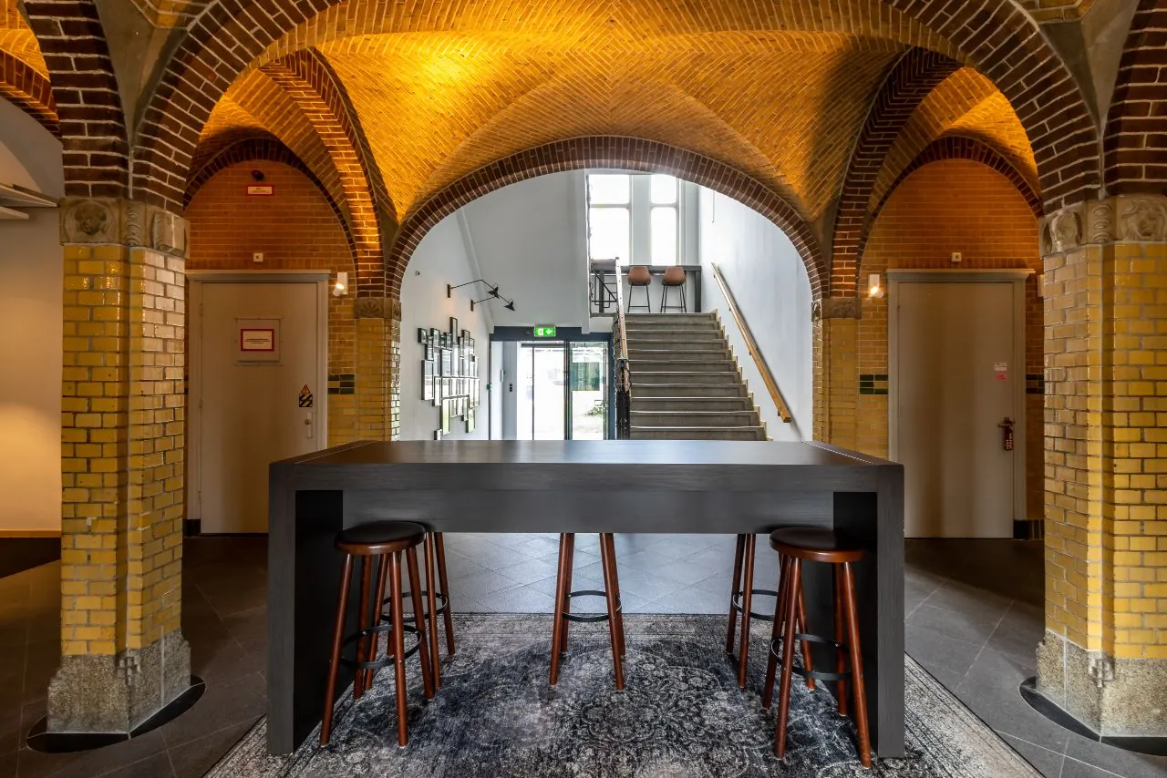 Hal met gewelfde bakstenen plafonds, een hoge tafel met krukken en een trap naar een lichtrijke bovenverdieping aan de Lage Naarderweg.